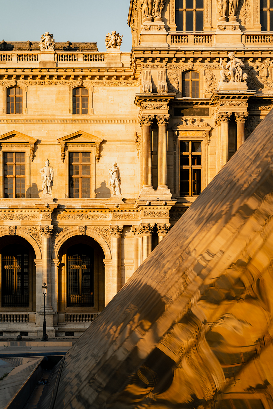 Golden hour detail of the Louvre façade with statues and arches, reflected in the glass pyramid in Paris, France