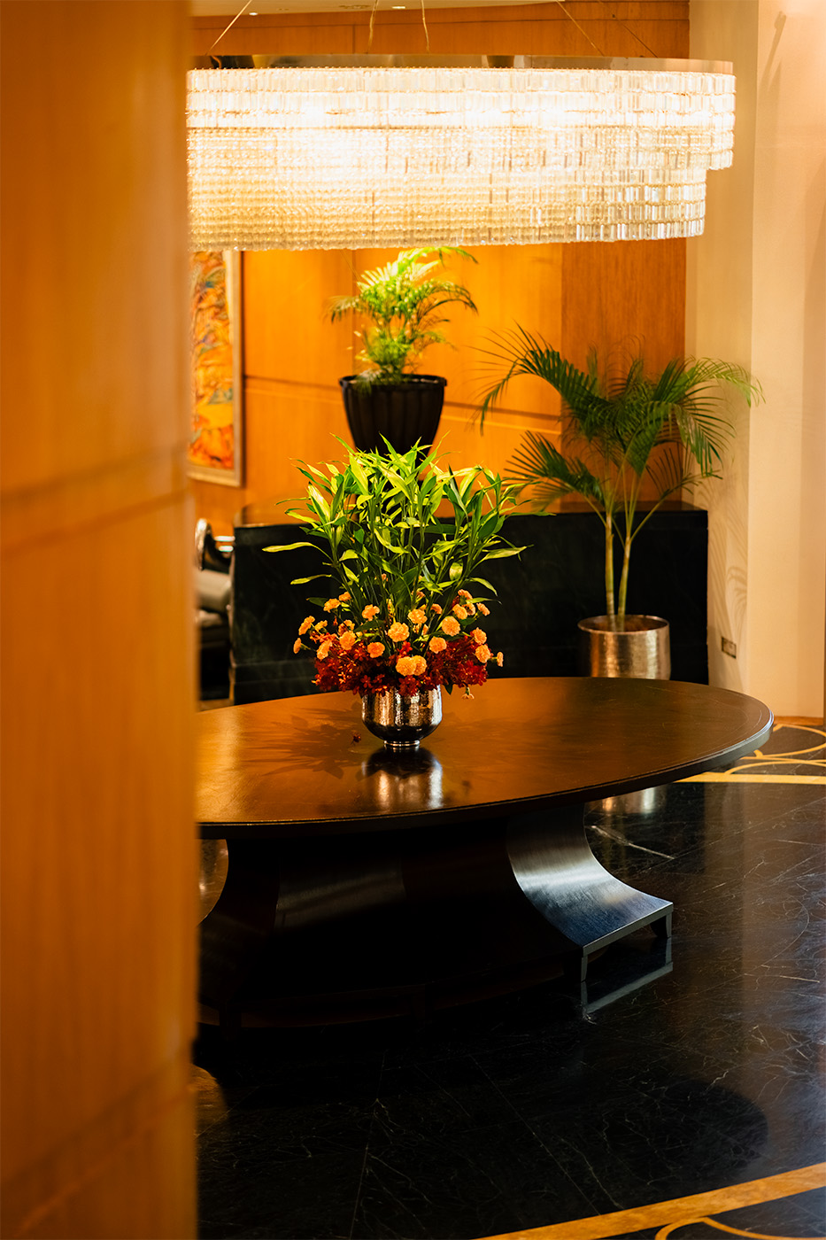 Round lobby table with a vibrant floral arrangement beneath a large crystal chandelier, warm wood-paneled background and indoor palms.
