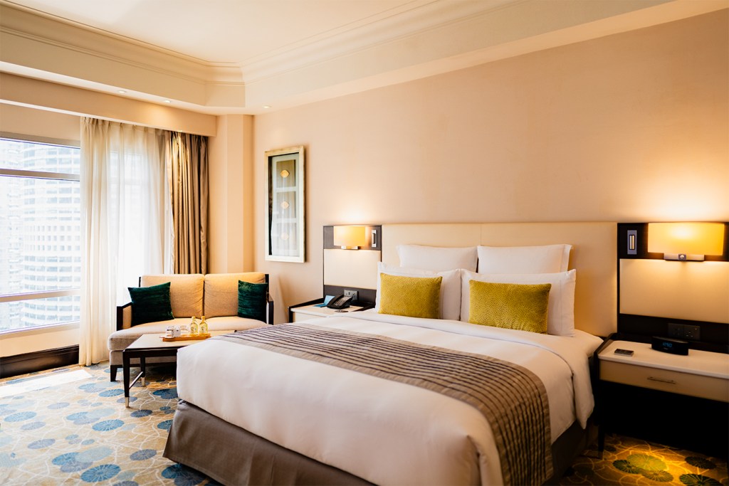 Bright hotel room with a king bed and green accent pillows, sofa seating by large windows overlooking the city.