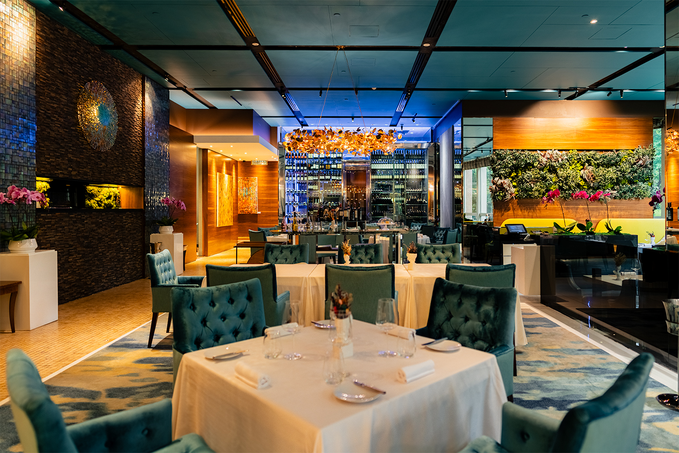 Elegant restaurant dining room with teal tufted chairs and white tablecloths, bar backdrop and a floral feature wall under warm lighting.