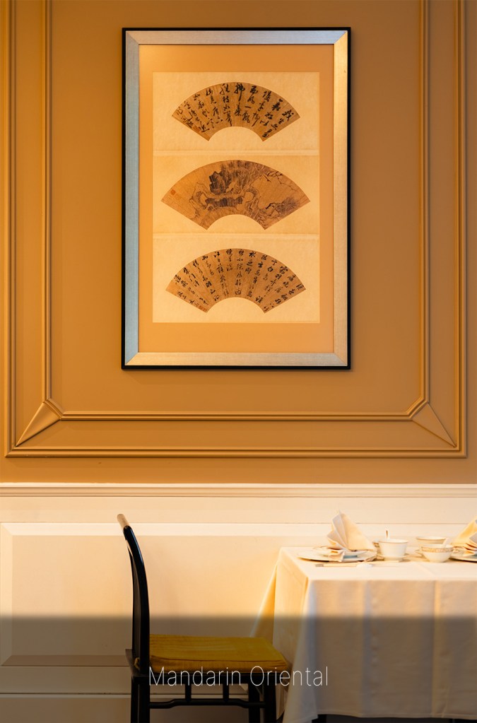 Framed fan artwork on a paneled wall above a dining table set with white linens, with a chair in the foreground.