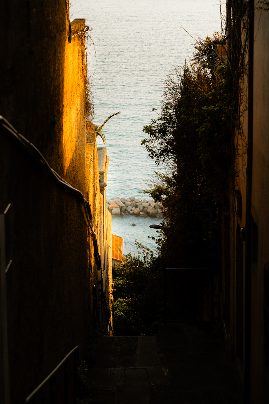 Narrow sunlit alley on a Marseille hillside opening onto the sea, with warm golden walls framing the water and rocks below