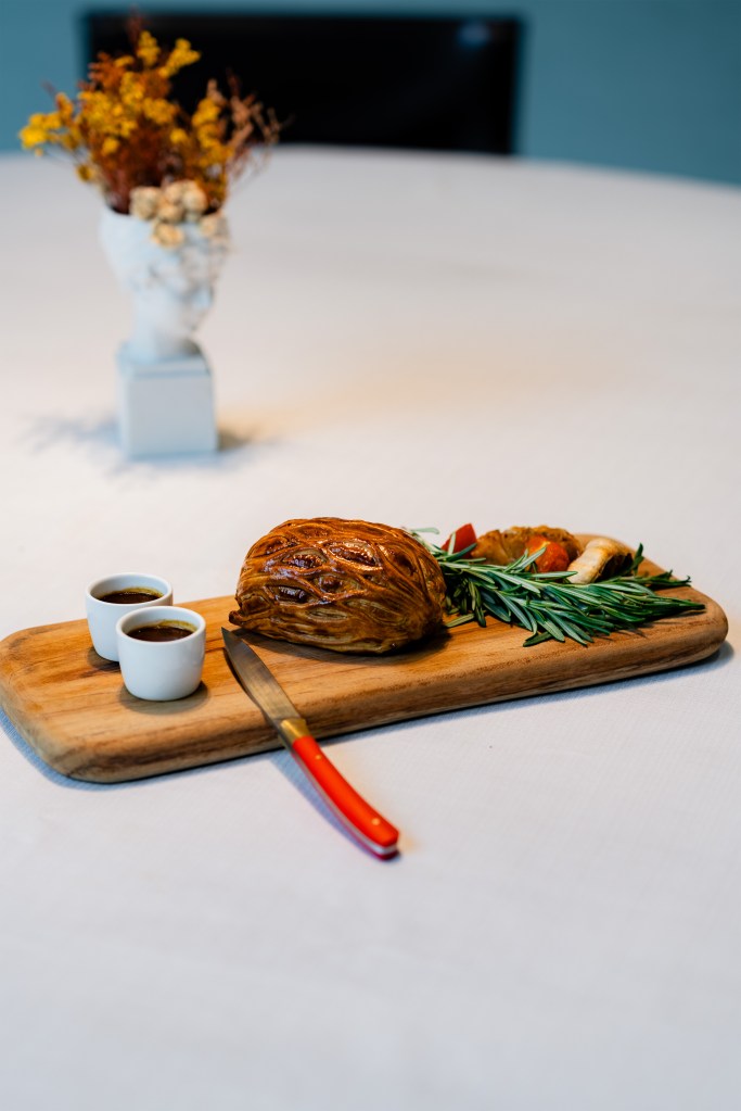 Roast dish on a wooden board with rosemary, sauce cups, and a red-handled knife on a white tablecloth, styled for service.