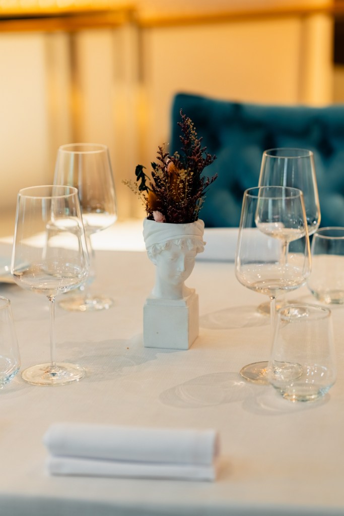 Table setting with wine glasses surrounding a classical bust vase filled with dried flowers, soft warm lighting and teal seating in the background.