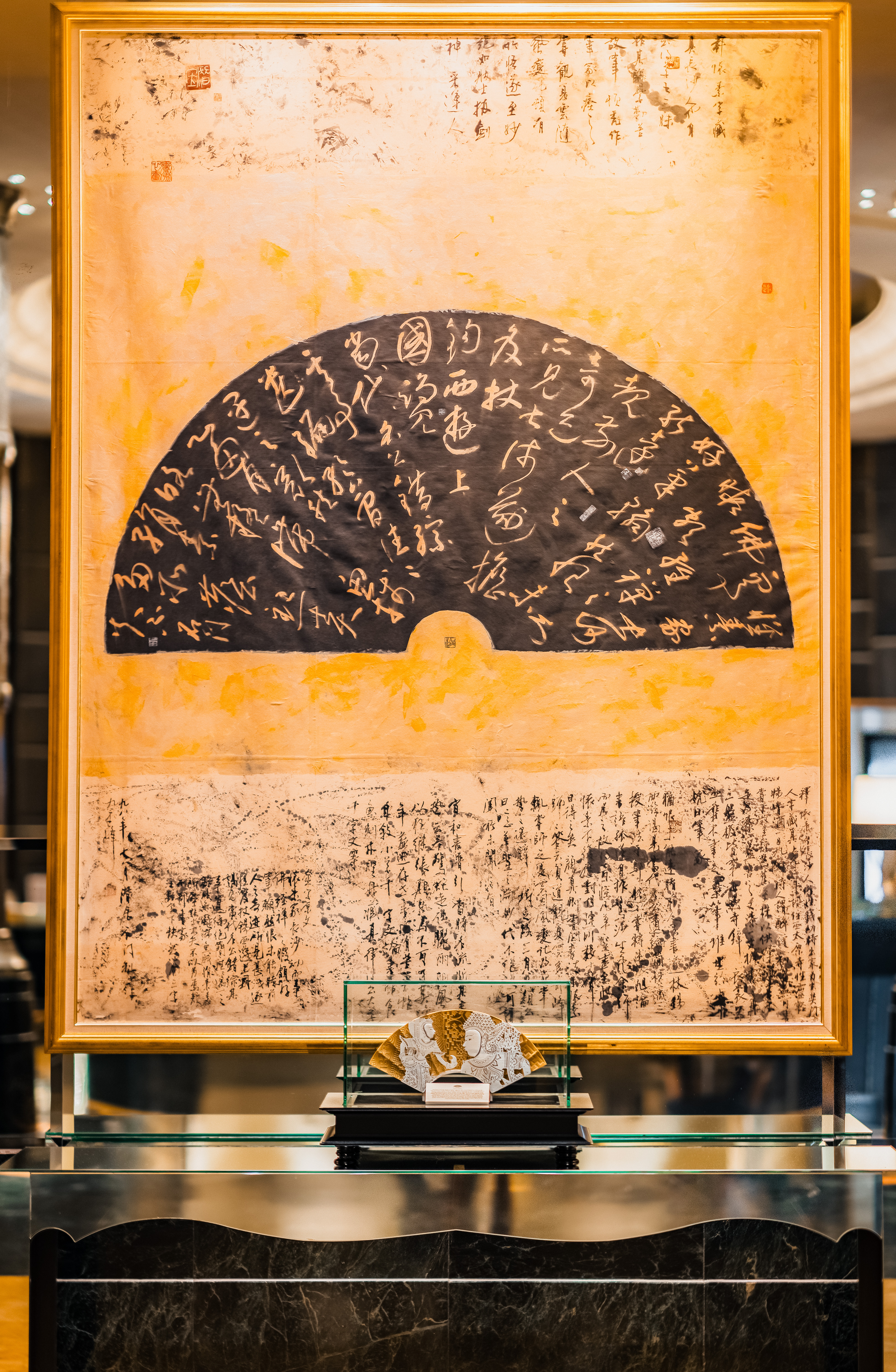 Large framed calligraphy artwork featuring a fan motif displayed above a console with a decorative fan sculpture in the foreground.