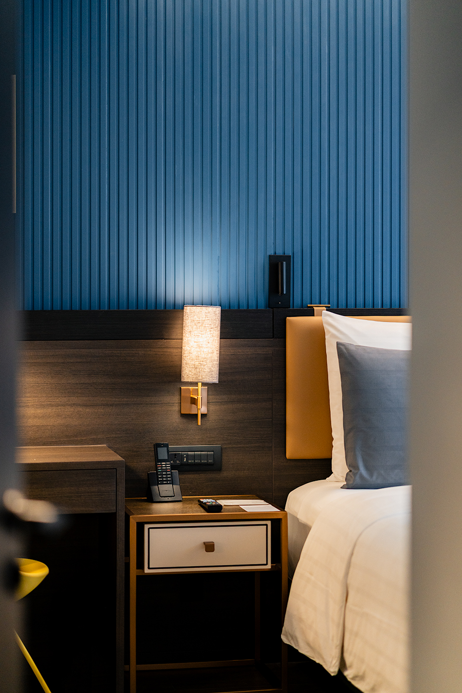 Bedroom corner detail at Sindhorn Midtown with a blue ribbed wall, warm wall sconce, bedside table, and crisp white bedding