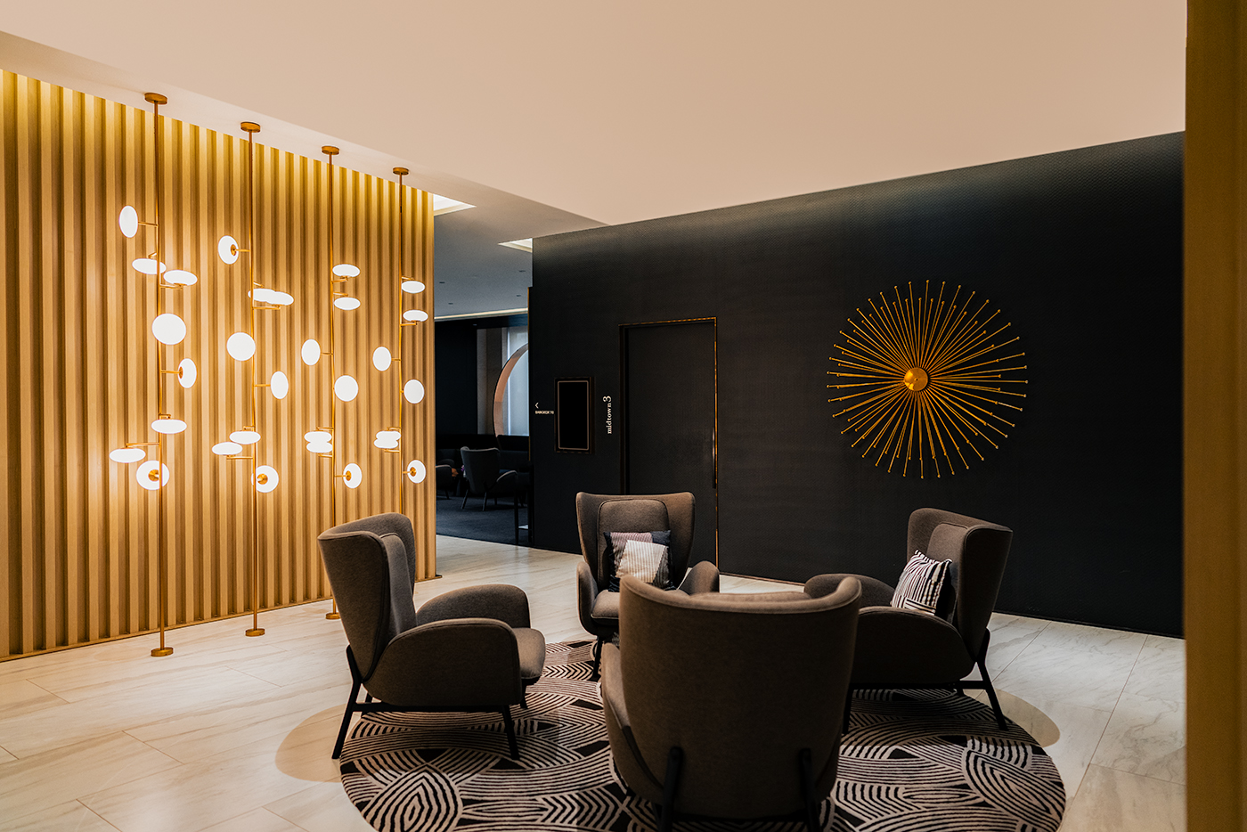 Modern lounge seating at Sindhorn Midtown with a gold-lit feature wall, circular sunburst wall sculpture, and four armchairs on a patterned rug