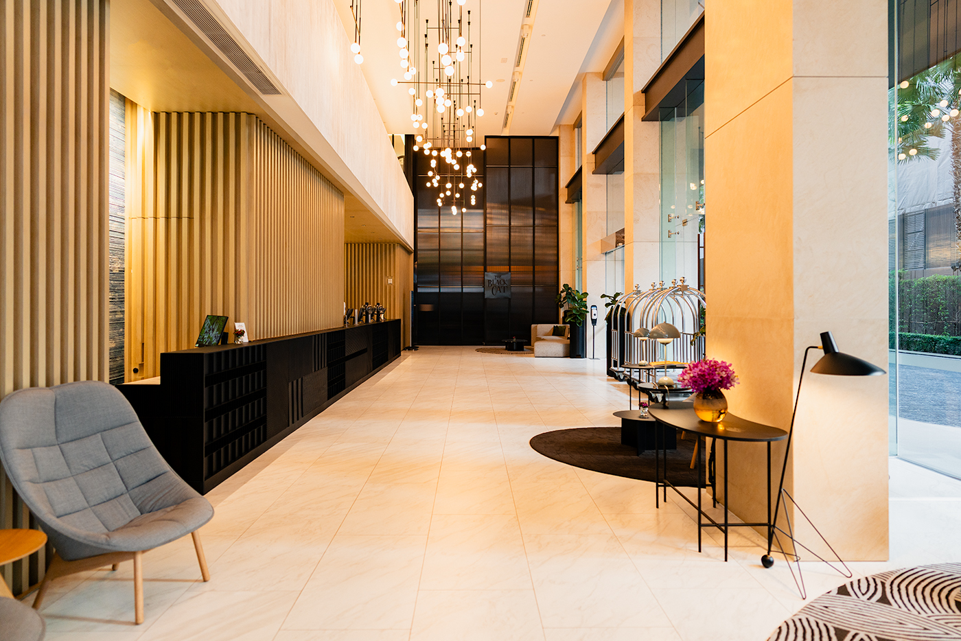 Bright hotel lobby at Sindhorn Midtown featuring a long black reception desk, hanging globe lights, marble floors, and luggage carts by the glass entrance