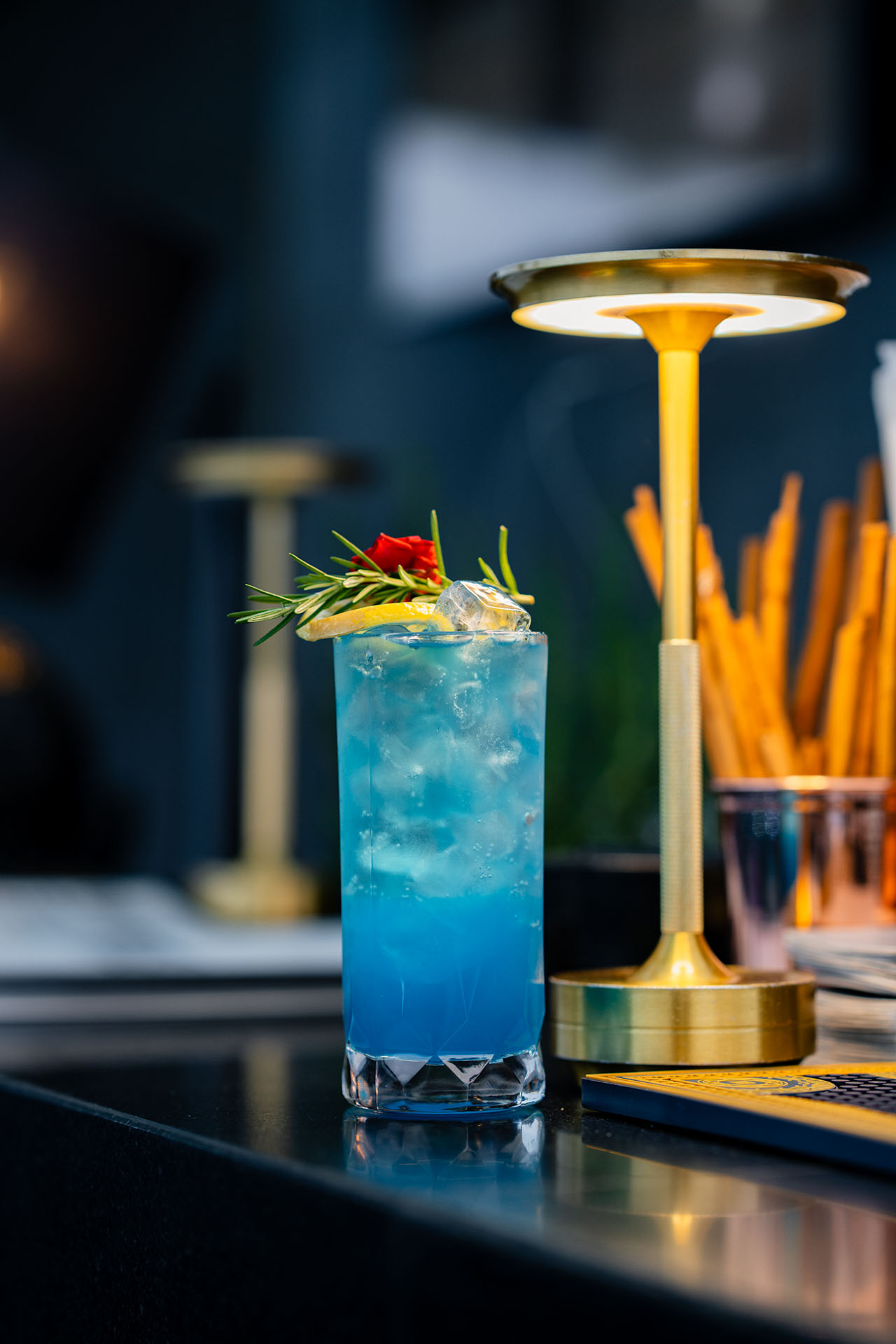 Blue signature cocktail garnished with rosemary and lemon on a dark bar counter with brass table lamp, Amanor Chiang Mai