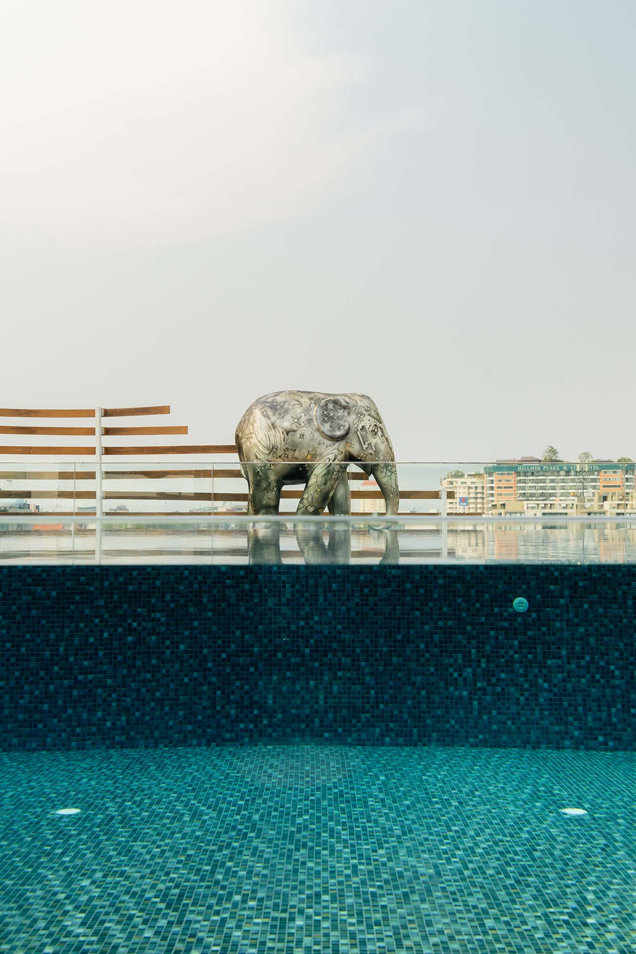 Rooftop infinity pool with engraved elephant sculpture at the water's edge against a hazy city skyline, Amanor Chiang Mai