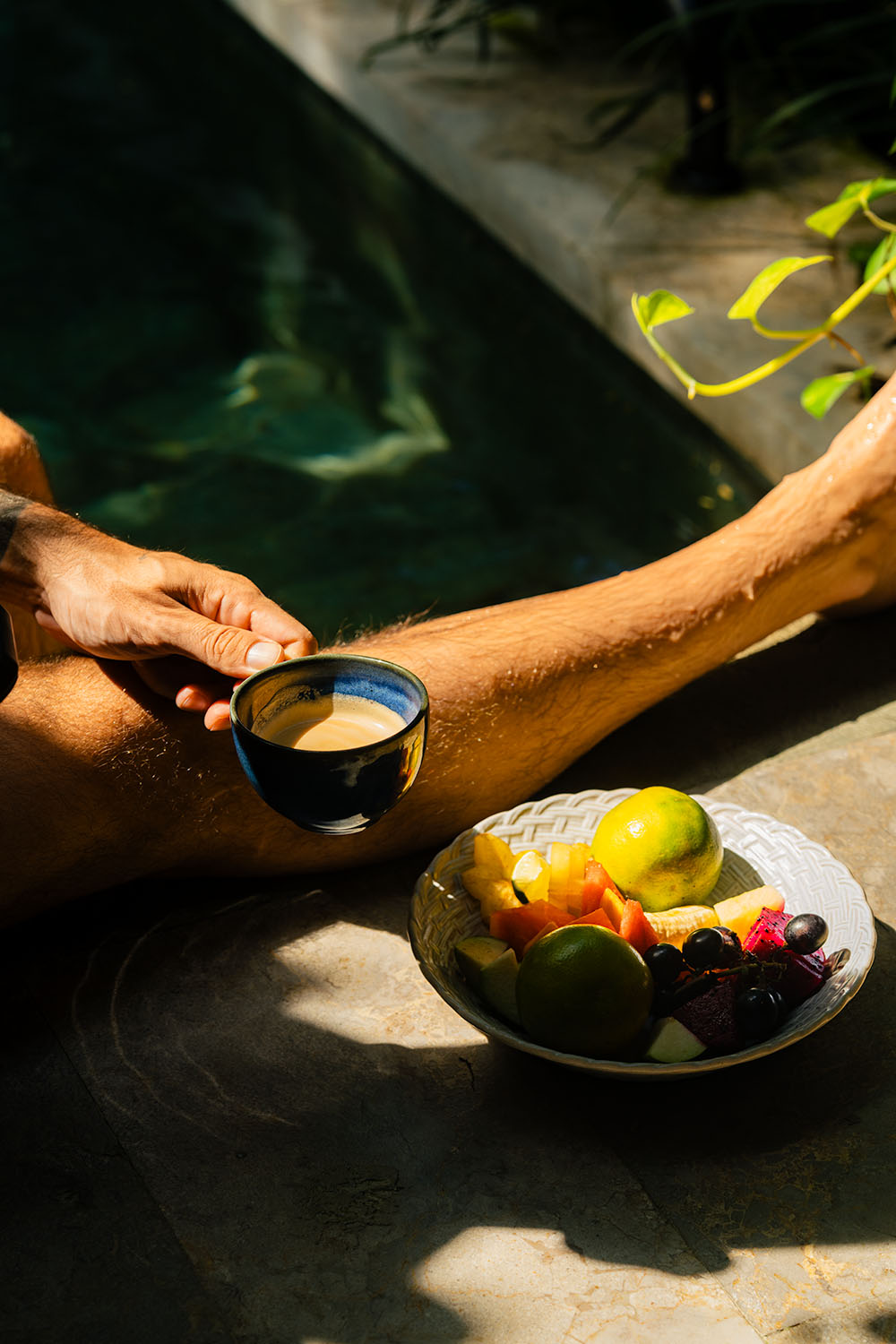 Guest holding an espresso cup poolside with a tropical fruit bowl in morning sun at Andaz Bali Sanur