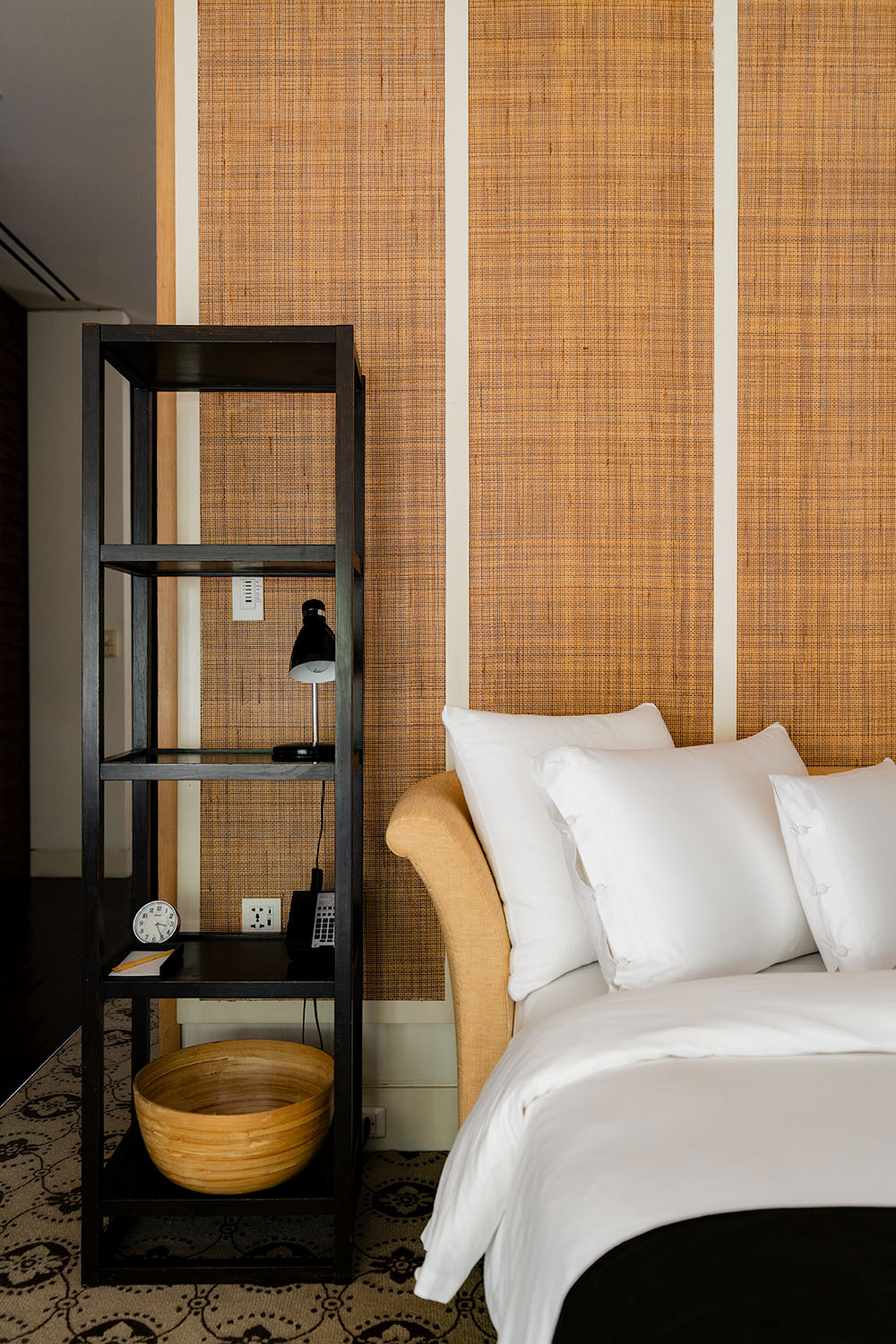Bedroom detail at Grand Hyatt Erawan Bangkok with woven rattan headboard, white linen, open-frame black shelf, and warm wall paneling