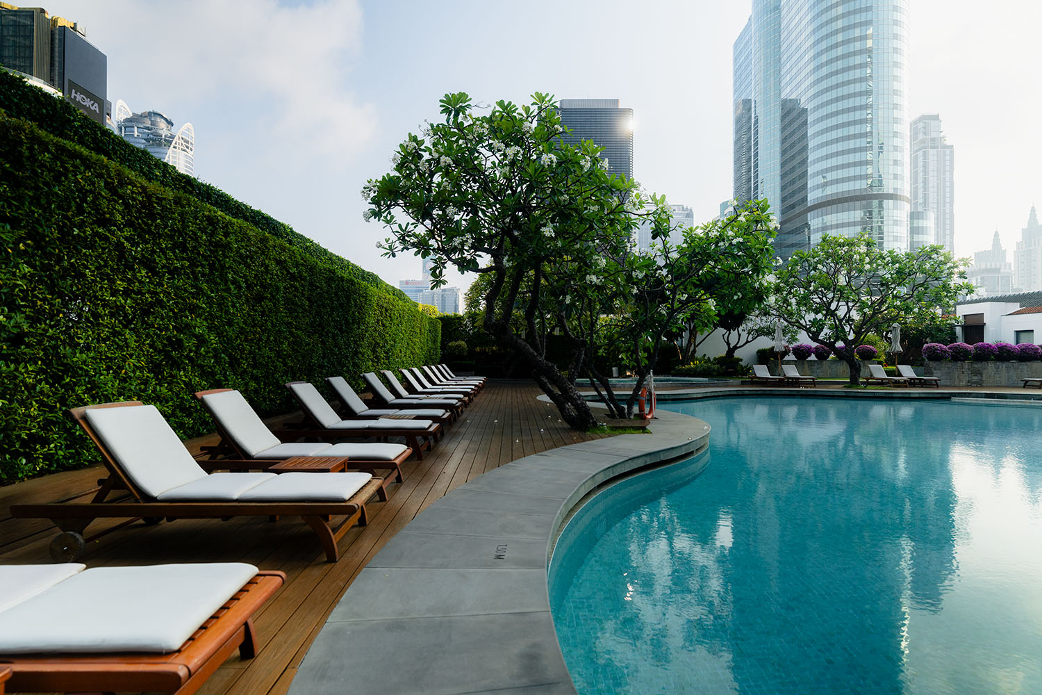 Outdoor pool at Grand Hyatt Erawan Bangkok with teak sun loungers, manicured hedges, frangipani trees, and Ratchaprasong skyline beyond