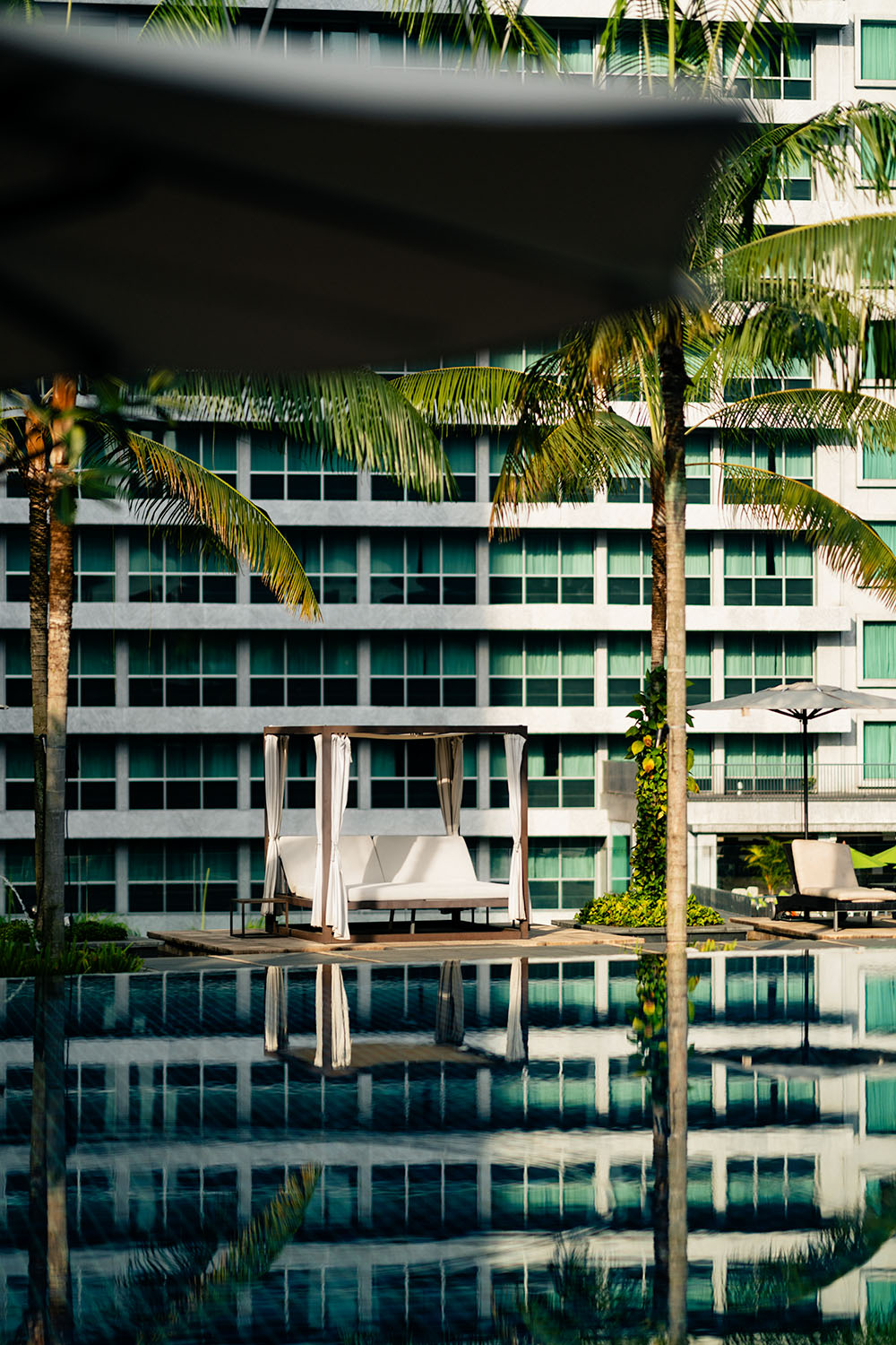 Poolside cabana with white curtains and sun loungers reflected in still water, palm trees framing Grand Hyatt Kuala Lumpur facade