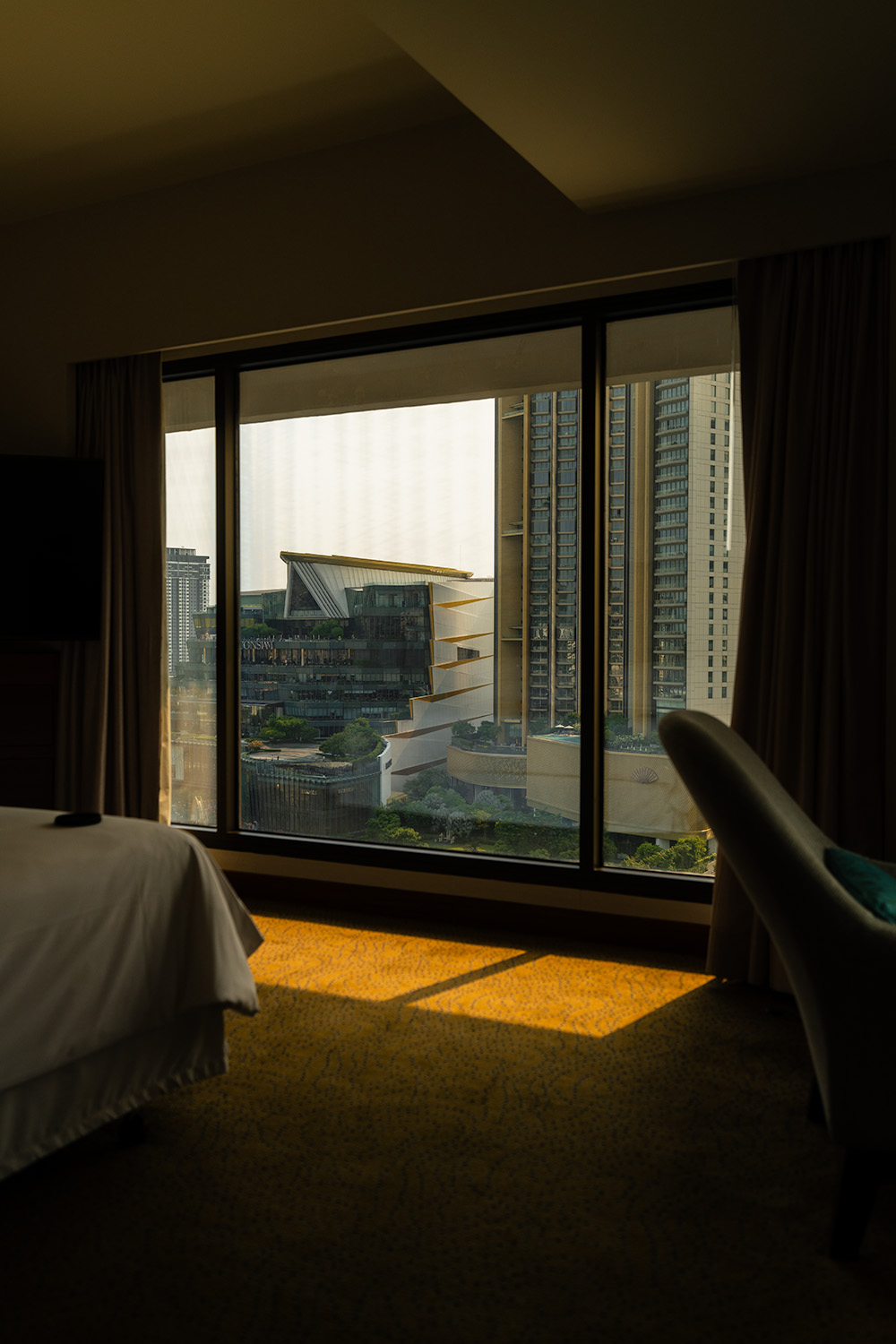 Hotel room at golden hour with floor-to-ceiling Bangkok city view and sunlight pooling on carpet, Sheraton Royal Orchid