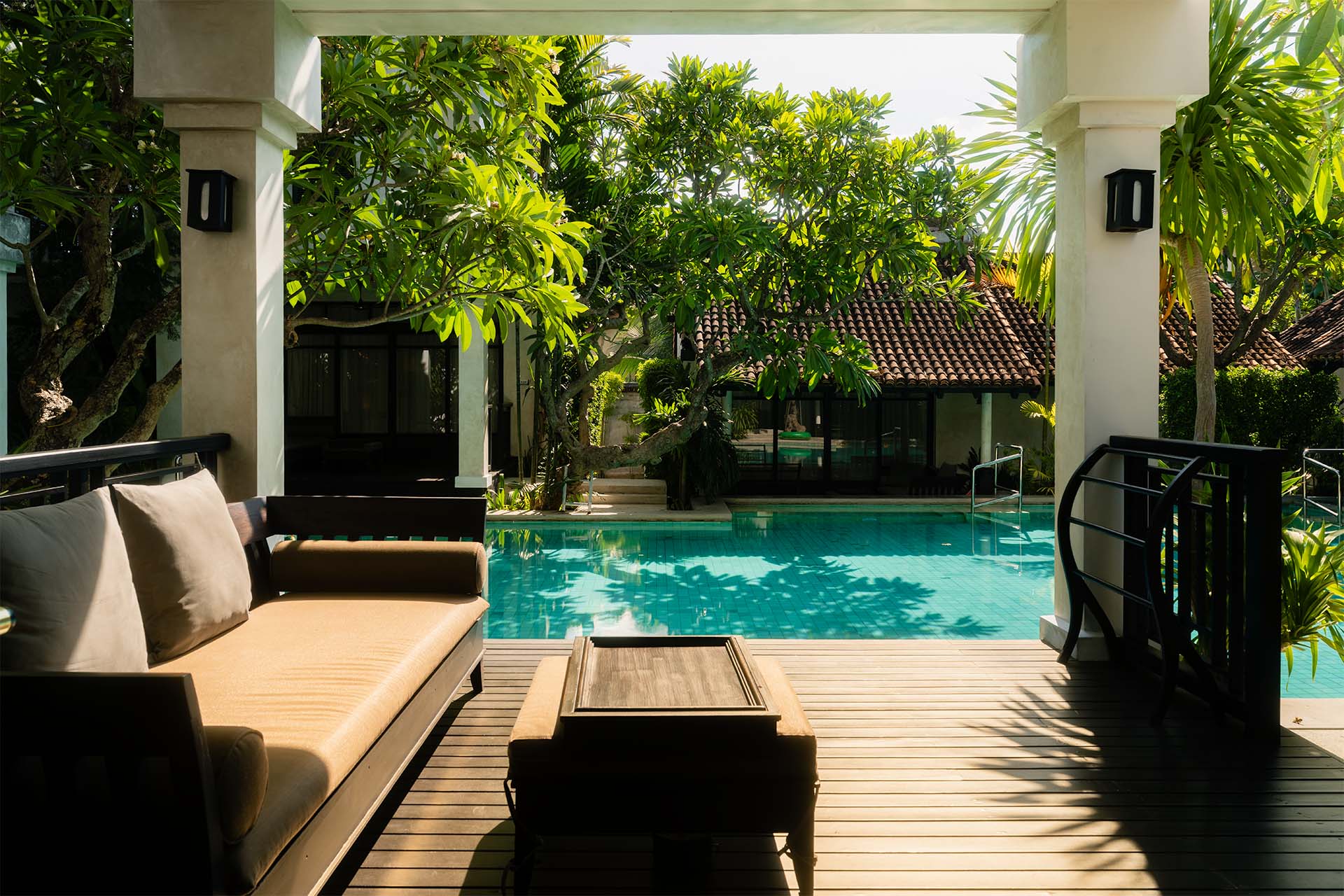 Shaded pool terrace at The Lamai Koh Samui with teak daybed, frangipani trees, and turquoise pool reflecting tropical garden