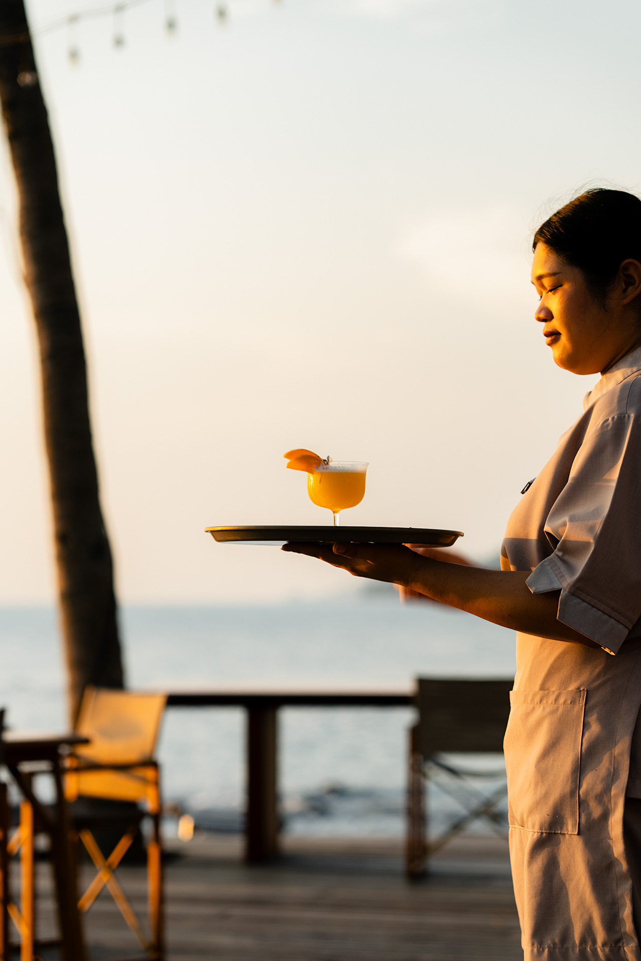Server carrying a golden cocktail on a tray at golden hour on the beachfront terrace of The Retreat Koh Chang, Thailand