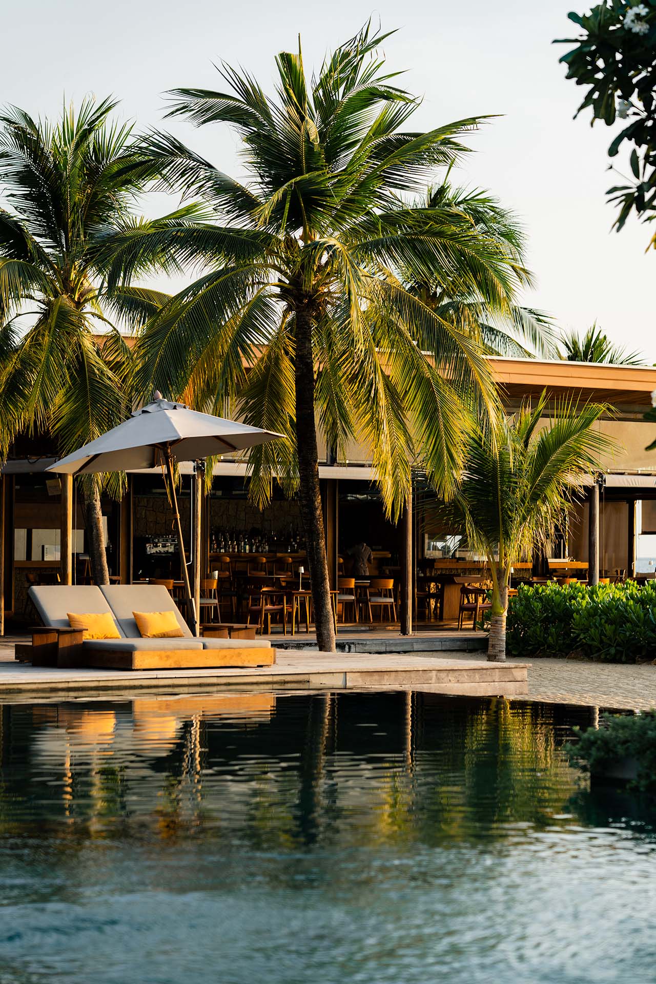 The Retreat Koh Chang beachfront pool at golden hour with yellow-cushioned daybeds, palm trees, and open-air beach bar reflected in still water