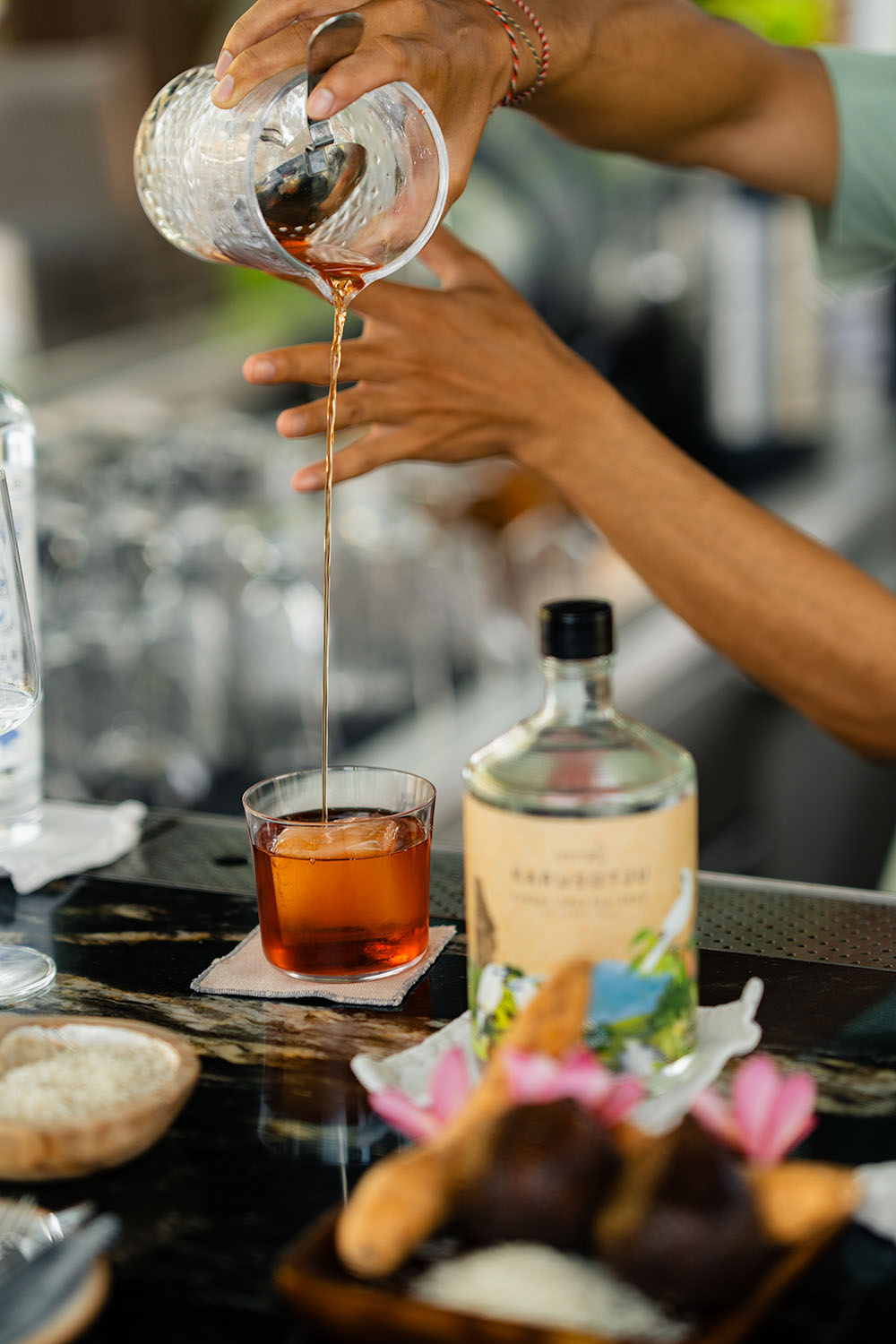 Bartender pouring a spirit over ice into a rocks glass at Umana Bali, with a botanically illustrated bottle and lotus flower on the bar